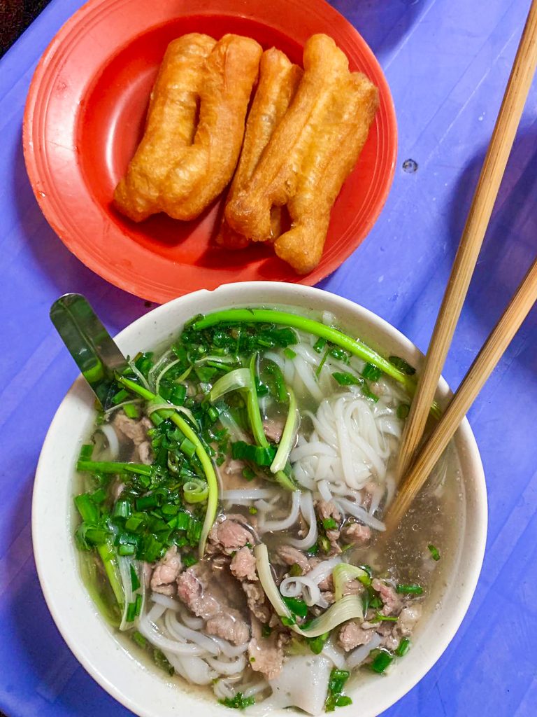 V miske vietnamska polievka Pho (ryžové rezance, hovädzie mäso na tenké pláty a veľa byliniek). V nej čínske paličky a lyžica a nad tým v miske sú nejaké vyprážané tyčinky