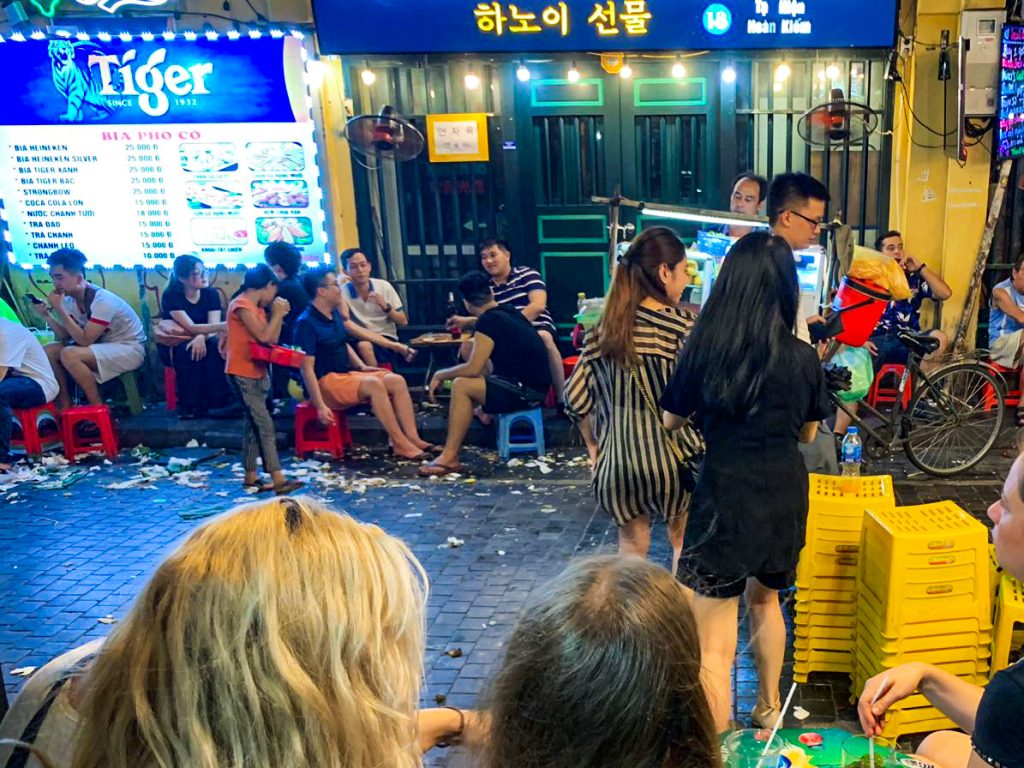 Ulička s barmi v centre Vietnamu, kde ľudia sedia na malých plastových stoličkách a okolo nich sú samé smeti