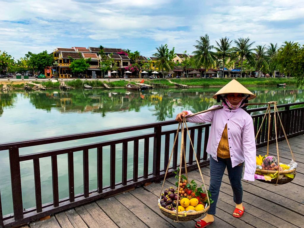 Vietnamka s typickým trojuholníkovým klobúkom na moste v Hoi An drži drevenú palicu na ramene a na jej oboch koncoch má zavesené veľké misy s ovocím na predaj. V pozadí rieka s ľoďkami, palmy a domy