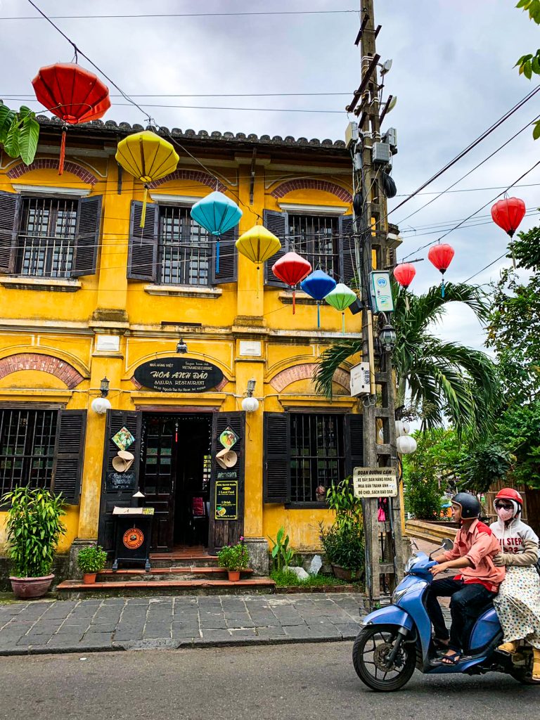 Sýto žltá budova v uliciach mesta Hoi An, pred ňou idú dvaja Vietnamci na motorke a hore je vidieť rôzno farebné lampióny povešané cez nad cestou