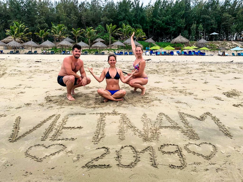 Ja, Miška a Igor na pláži b plavkách pri nápise v piesku Vietnam 2019 s dvoma srdiečkami po oboch stranách nápisu