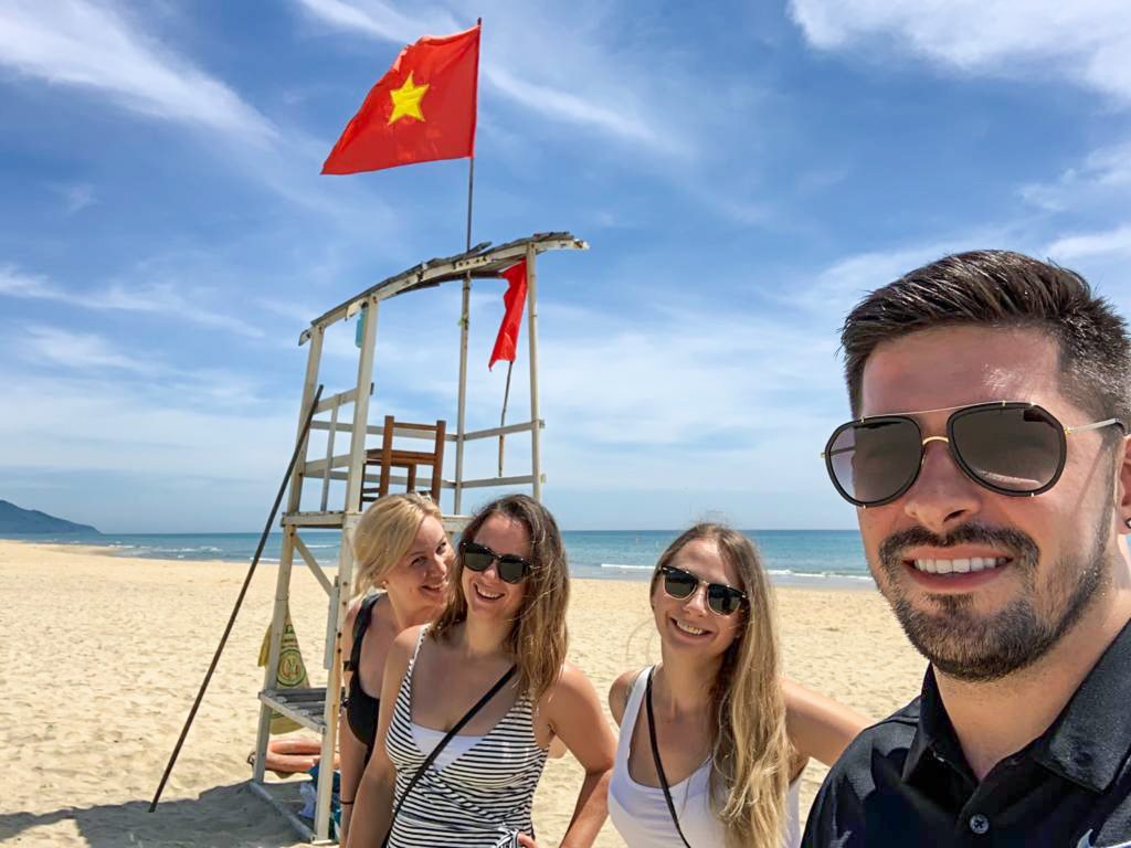 Selfie nás štyroch na prázdnej pláži v pozadí plavčícka veža, na ktorej veje vietnamská vlajka