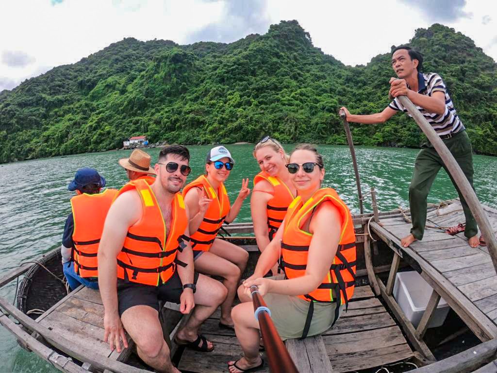 My štyria v oranžových záchraných vestách v malej drevenej loďke v zálive poháňanej chudým Vietnamcom pomocou veľkách pádiel