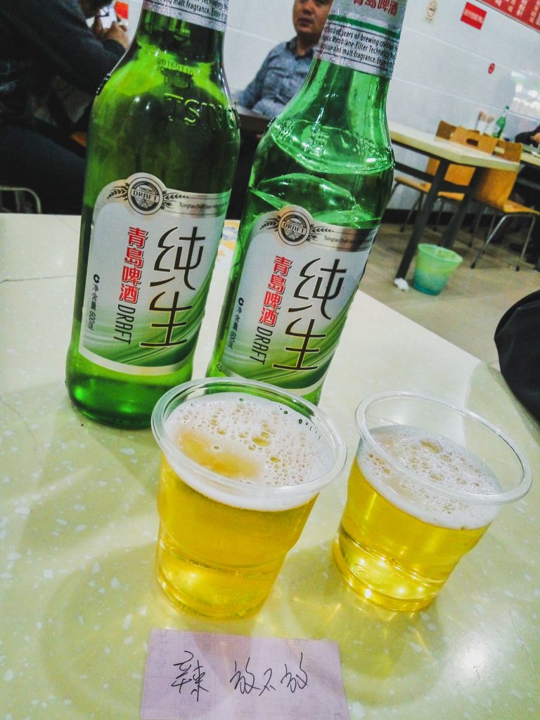 Dve fľaše čínskeho piva na stole, dva plastové poháre plné piva a pred tým položený malý fialový papierok s nejakými čínskymi znakmi