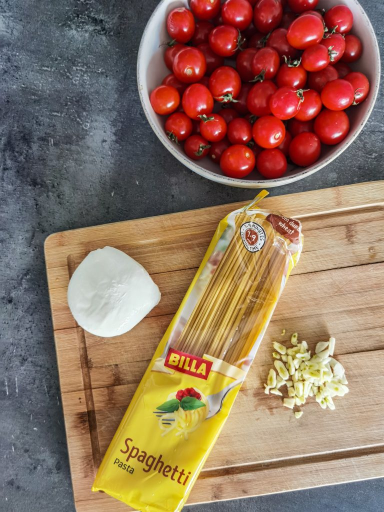 Cherry rajčiny v miske. Na krájacej doske guľka mozzarely, nasekaný cesnak a zabalené špagety.