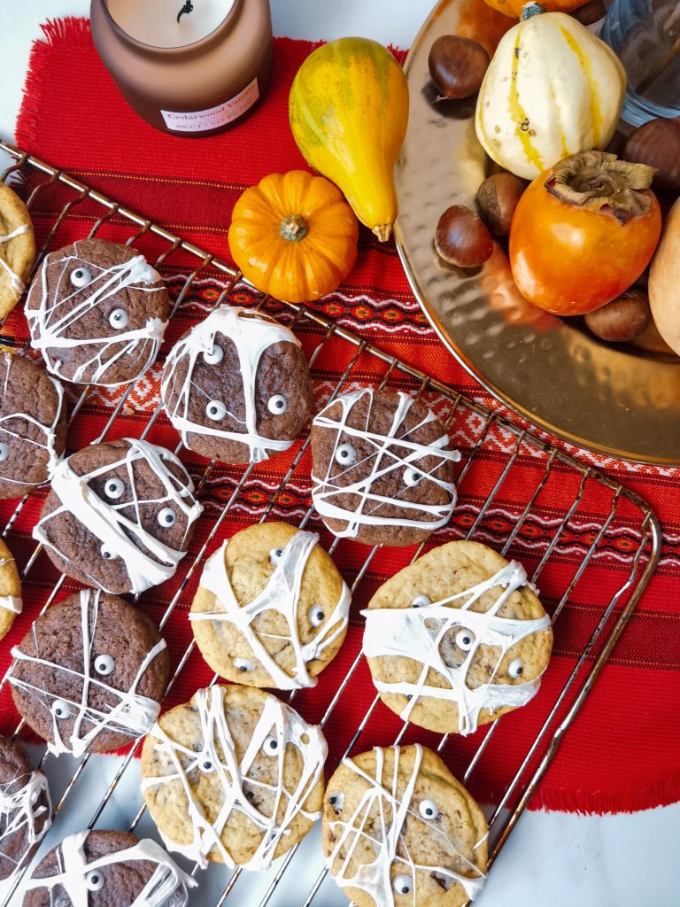 Halloweenske cookies ozobené cukrovými očami a múmiovskou bielou sieťou sú naukladané na mriežke na červenom obruse a v pozadí vidieť malé tekvice
