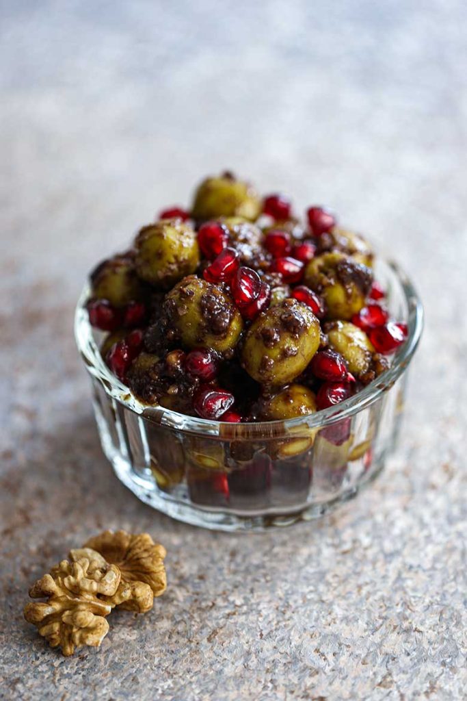 Nakladané olivy v priesvitnej miske posiate jadierkami z granátového jablka, vpredu vidieť vlašský orech