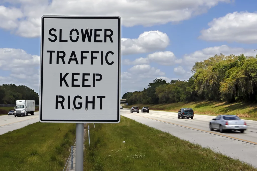 V popredí biela značka s nápisom "Slower traffic keep right" a v pozadí po oboch stranách jednosmerné pruhy diaľnice s autami v pohybe