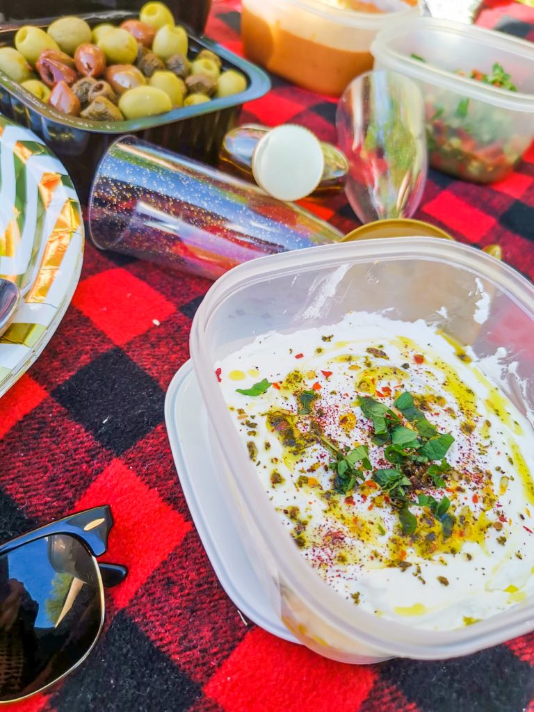 Labneh v miske na pikniku, v pozadí poháre na šampanské, mix olív a izraelský šalát