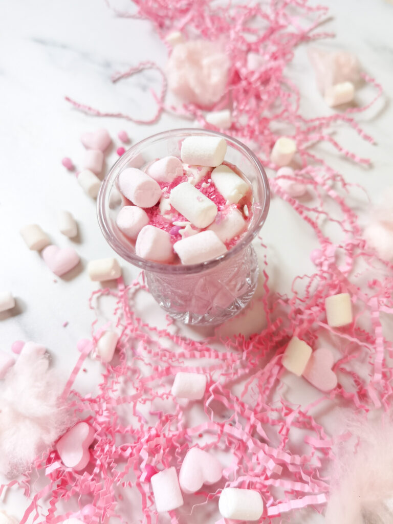 ružovú horúca čokoláda podávaná v malom priehľadnom pohári na vrchu zasiata mini marshmallows a ružovými trblietkami, okolo pohádzané ružové papieriky, marshmallows a cukrové srdiečka