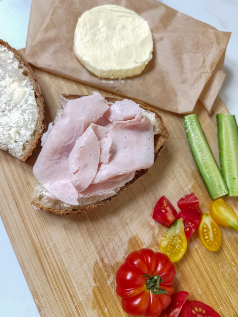 kúsok chlebu so šunkou, rajčinami, v pozadí domáce maslo